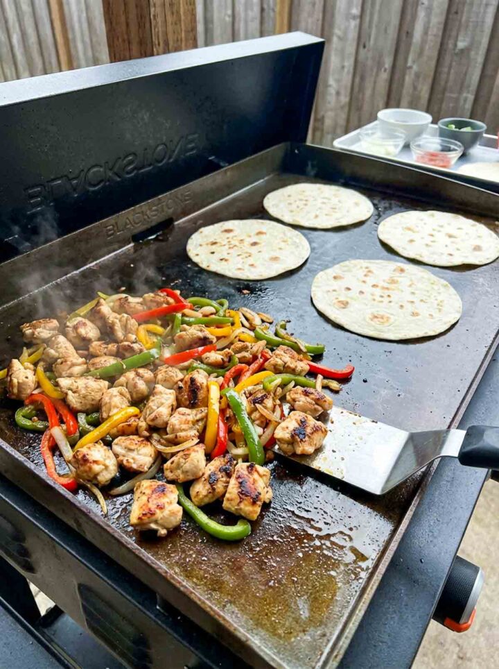 Chicken fajitas with peppers and tortillas cooking on a Blackstone griddle with light char and steam.