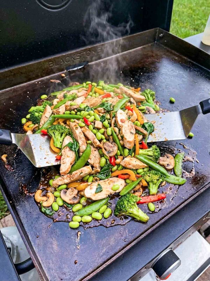 Chicken stir fry with broccoli, snap peas, edamame, and peppers cooking on a Blackstone griddle with steam.