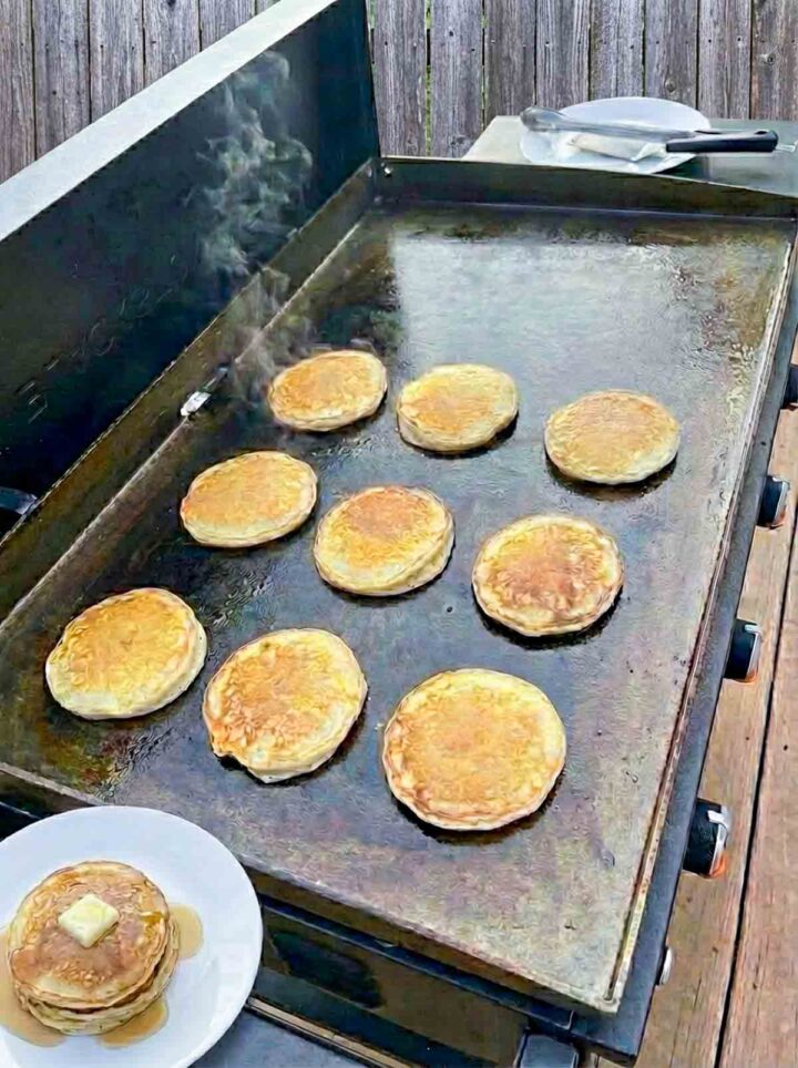Cottage cheese protein pancakes cooking on a Blackstone griddle with a stacked serving topped with butter and syrup on the side.