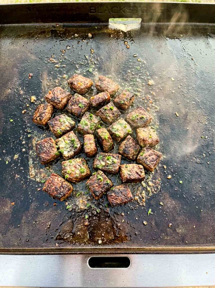 Sizzling garlic butter steak bites cooking on a Blackstone griddle, seared on all sides and topped with herbs, surrounded by melted butter and steam.