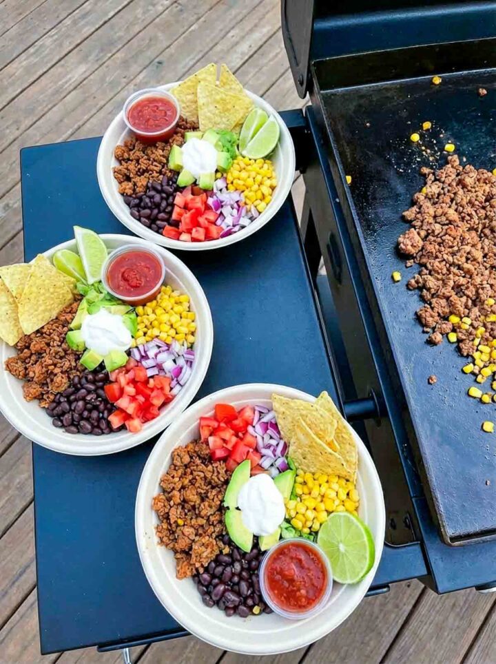 Ground turkey taco bowls with rice, beans, corn, avocado, and toppings beside a Blackstone griddle.