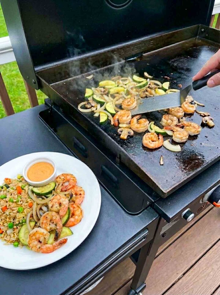 Hibachi shrimp with zucchini and onions cooking on a Blackstone griddle with a plated serving of fried rice and sauce on the side.