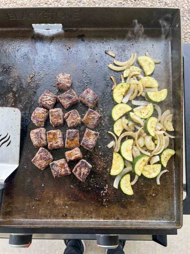 Hibachi steak cubes with zucchini and onions cooking on a Blackstone griddle in separate sections.