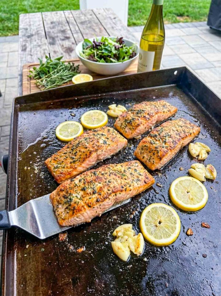 Lemon garlic salmon fillets cooking on a Blackstone griddle with butter, herbs, and lemon slices.