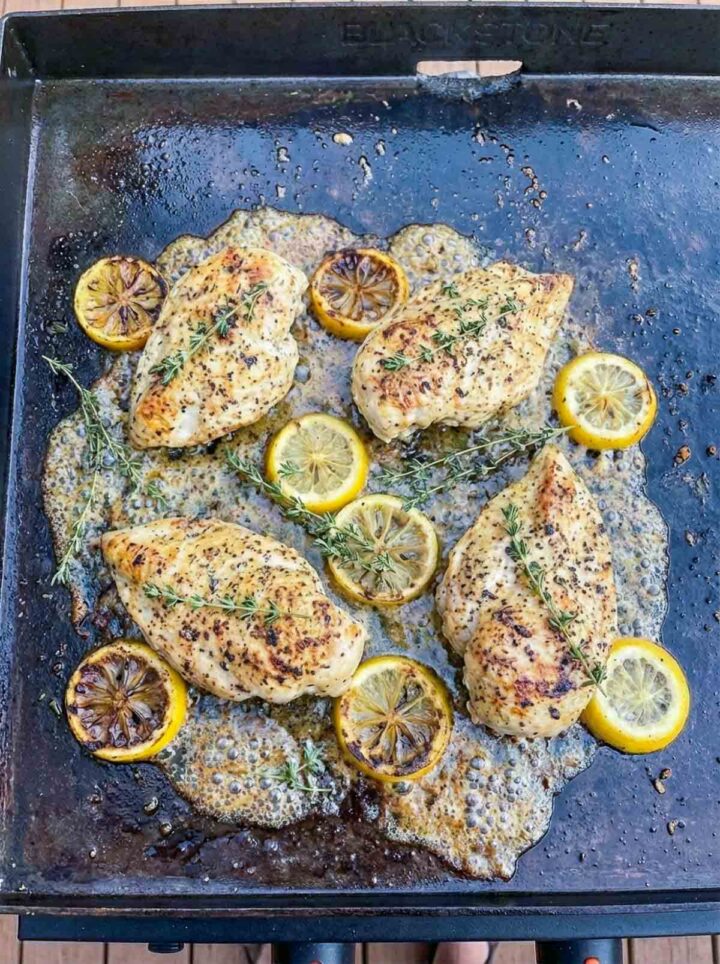 Lemon pepper chicken breasts cooking on a Blackstone griddle with butter, herbs, and caramelized lemon slices.