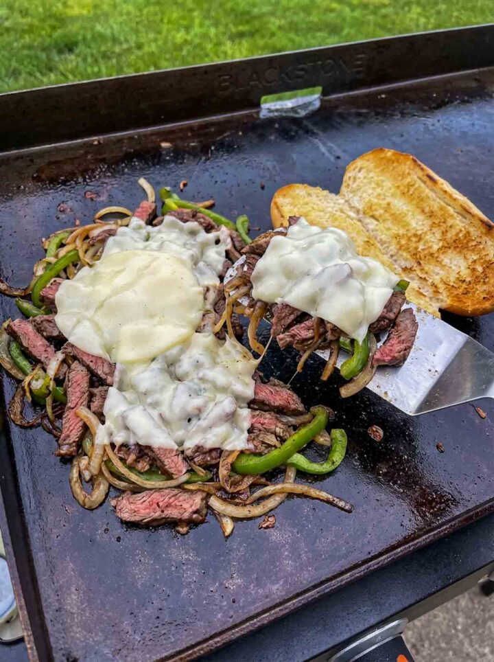 Philly cheesesteak filling with melted cheese, peppers, and onions on a Blackstone griddle with toasted roll.