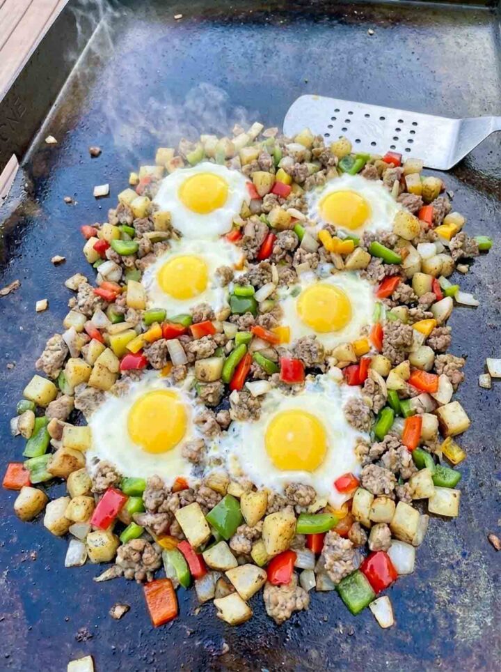 Turkey sausage breakfast hash with crispy potatoes, peppers, and six sunny side eggs cooking on a Blackstone griddle.
