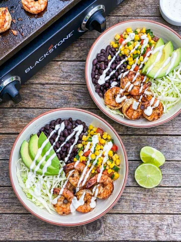 Shrimp burrito bowls with black beans, corn, avocado, and sauce near a Blackstone griddle.