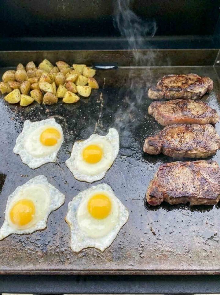 Steak and eggs cooking on a Blackstone griddle with crispy potatoes and steam rising.