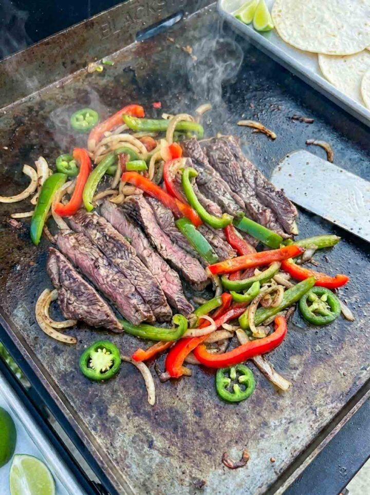Steak fajitas cooking on a Blackstone griddle with sliced beef, sautéed bell peppers, onions, and jalapeños, with tortillas and lime wedges nearby.
