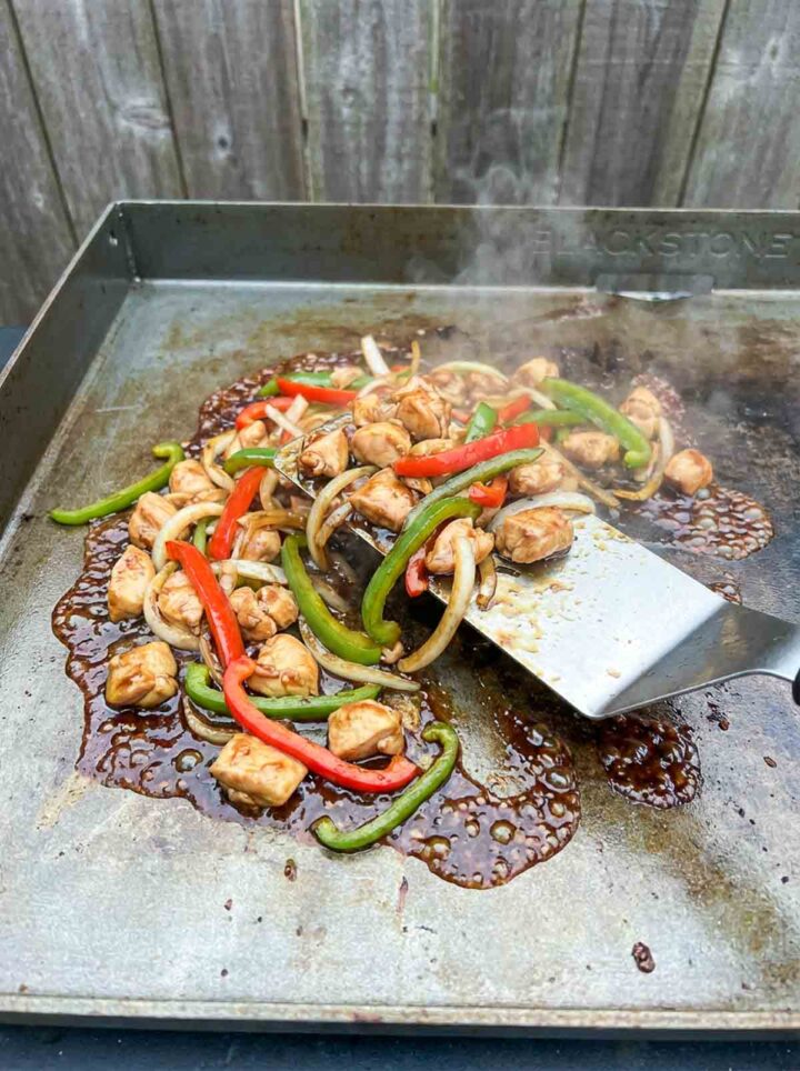 Teriyaki chicken with peppers and onions cooking on a Blackstone griddle with glossy sauce and steam.