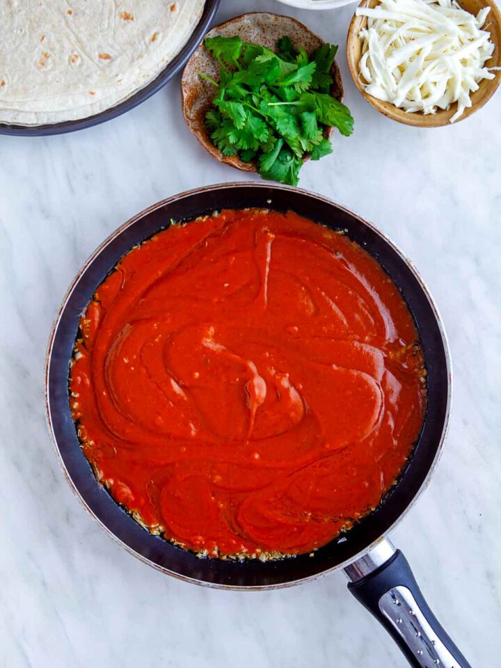 Red enchilada sauce cooking in a skillet with tortillas, cilantro, and shredded cheese nearby.