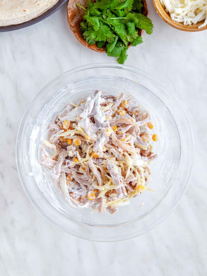 Shredded chicken mixed with corn, cheese, and creamy sauce in a glass bowl for enchilada filling.