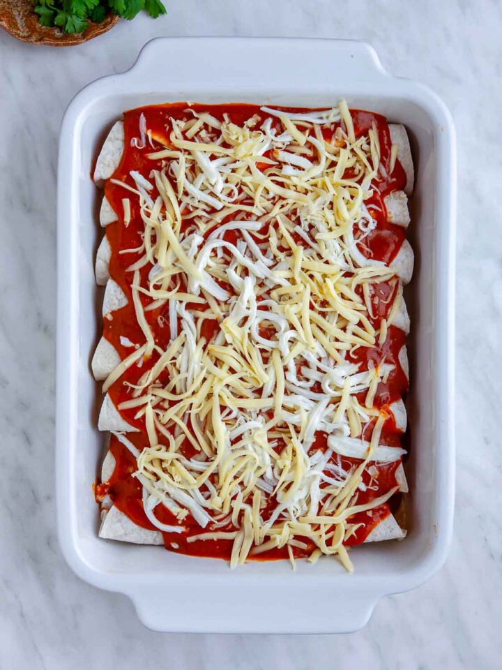 Chicken enchiladas in a baking dish topped with red sauce and shredded cheese before baking.