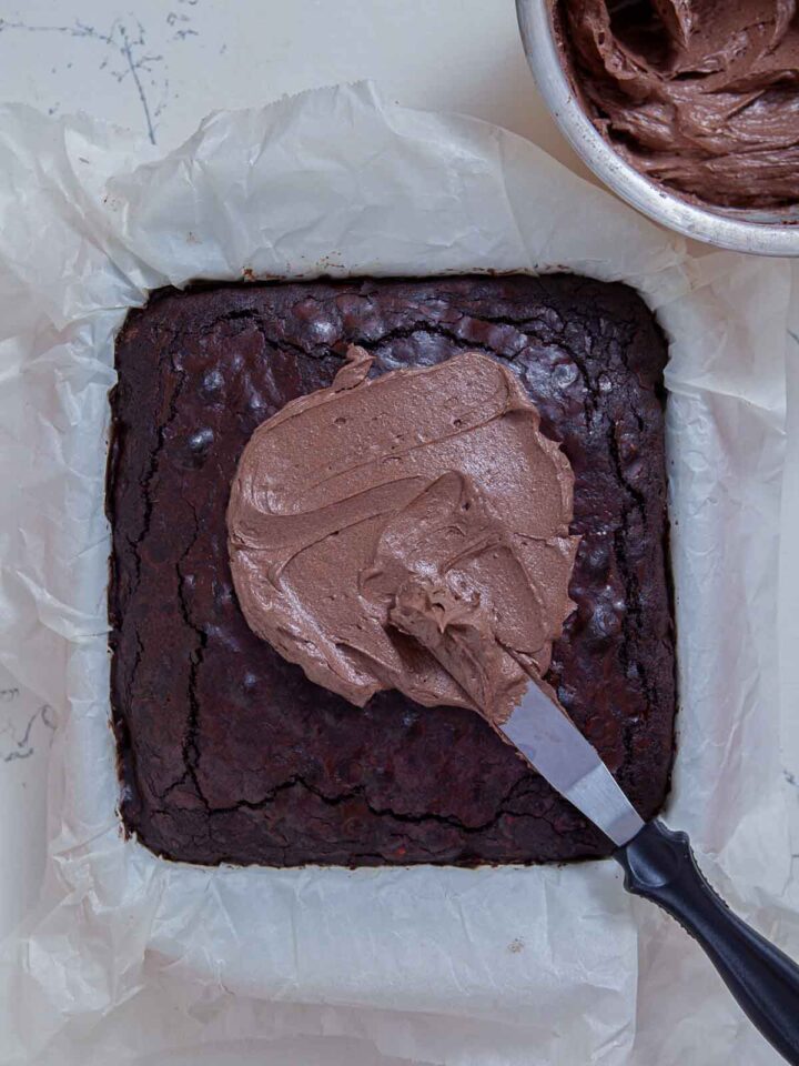 Chocolate frosting being spread over freshly baked brownies in a pan.