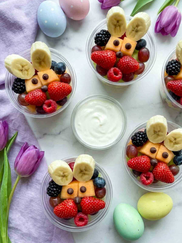 Clear snack cups filled with colorful fruit arranged like bunny faces using banana slice ears, chocolate chip eyes, strawberries, grapes, and berries, served with a bowl of yogurt dip on a white marble surface with pastel Easter eggs and tulips.