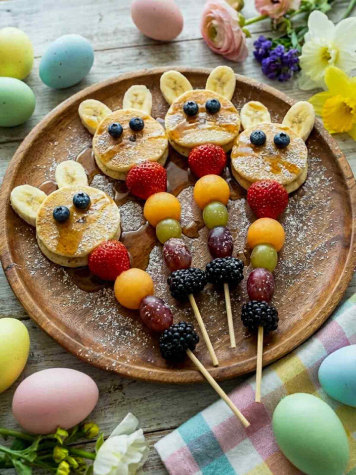 Easter bunny pancake stacks with banana ears and blueberry eyes served with colorful fresh fruit skewers on a wooden plate surrounded by pastel Easter eggs and spring flowers.