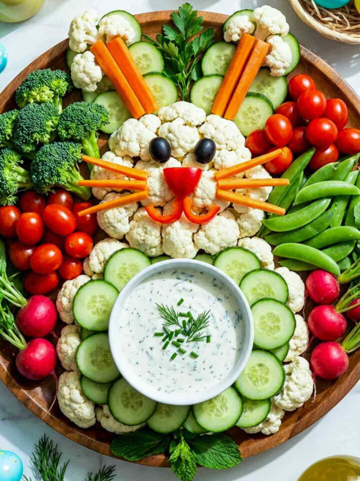 Overhead photo of a bunny-shaped veggie tray arranged on a round wooden platter. Cauliflower florets form the bunny face, with black olive eyes, a red radish nose, and carrot whiskers. Cucumber slices surround a bowl of ranch dip in the center, while broccoli, cherry tomatoes, snap peas, and radishes fill the rest of the tray. Pastel Easter eggs and spring flowers decorate the background.