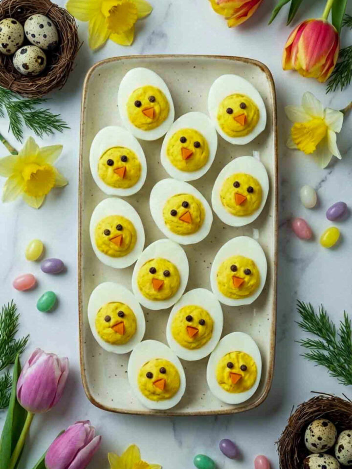 Top view of deviled eggs styled like baby chicks with carrot beaks and pepper eyes arranged on a rectangular serving tray surrounded by spring flowers and pastel Easter candy.