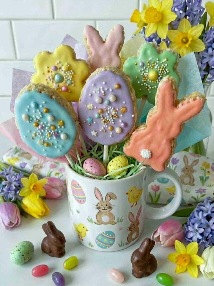 Pastel Easter egg and bunny shaped Rice Krispie treat pops on sticks arranged in a mug filled with decorative Easter grass, surrounded by spring flowers, chocolate bunny candies, and colorful jelly beans on a white marble surface.