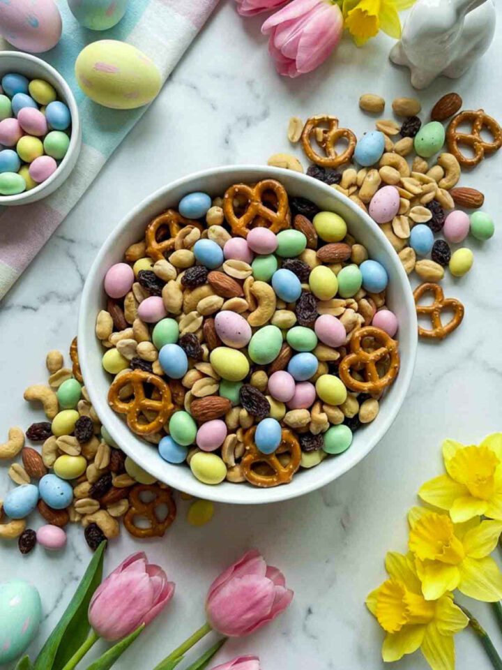 Easter trail mix with yogurt candy eggs, pretzels, nuts, and raisins served in a white bowl with pastel eggs and spring flowers.