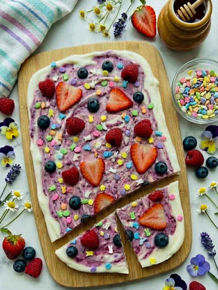 Top view of swirled frozen yogurt berry bark on a wooden board topped with strawberries, raspberries, blueberries, and pastel Easter sprinkles with small bowls of toppings around it.