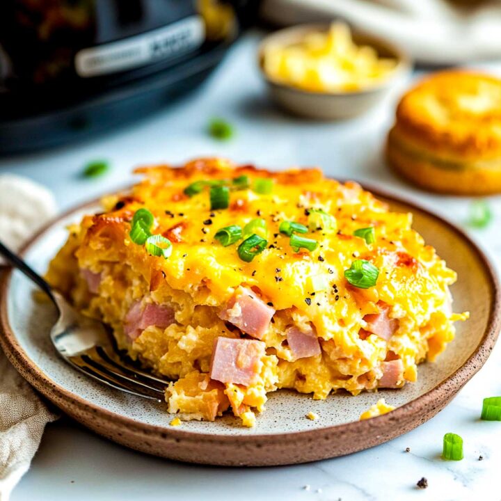 A slice of ham and cheese casserole served on a plate with crockpot in the background.