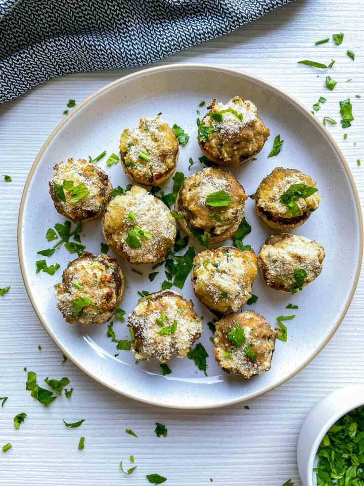 Plate of baked stuffed mushrooms topped with grated cheese and fresh parsley, served as a savory appetizer.