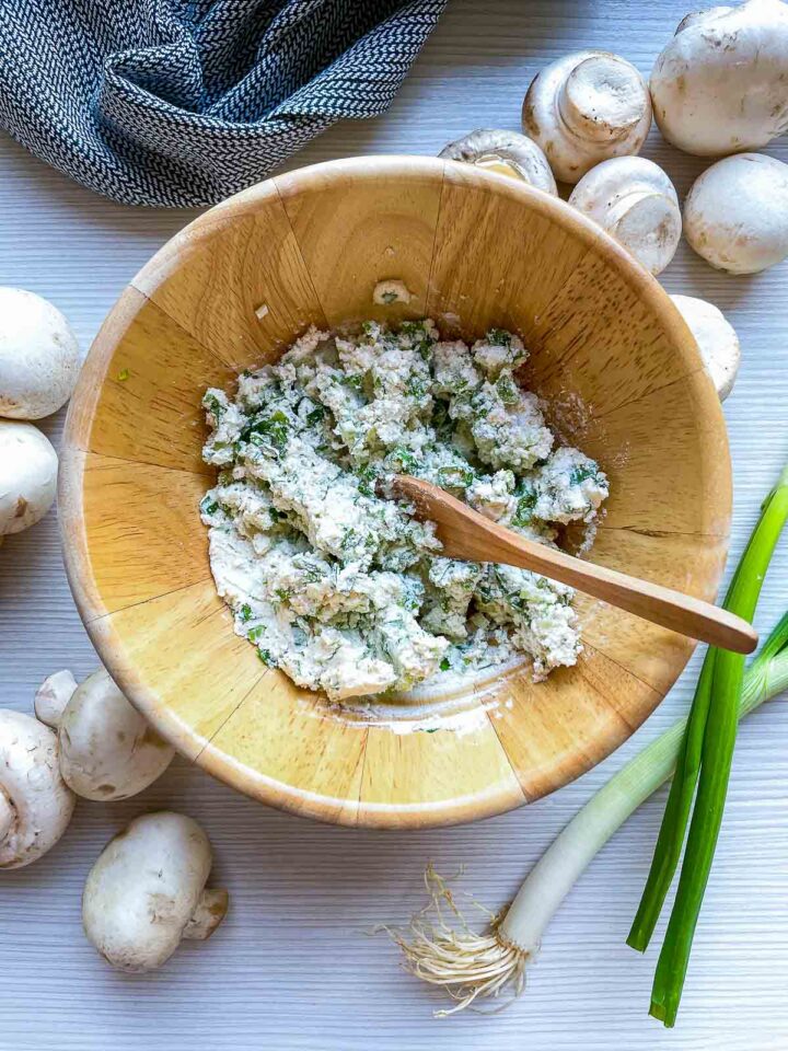 Wooden bowl with herbed cream cheese filling and spoon, surrounded by mushrooms and green onions.