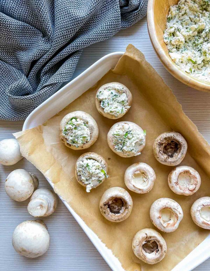 Raw mushroom caps filled with herbed cream cheese mixture arranged on a parchment-lined baking tray.