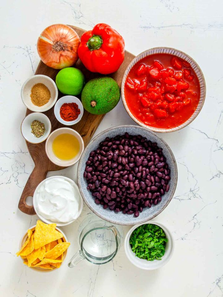 Ingredients for black bean nacho soup including beans, tomatoes, avocado, spices, onion, lime, tortilla chips, and sour cream.