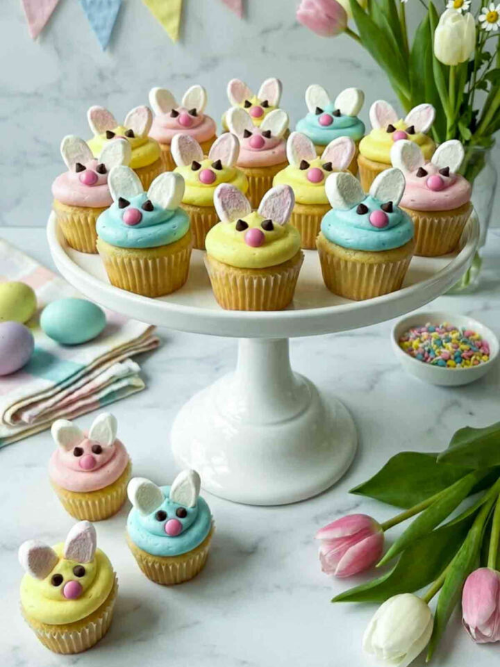 Mini bunny cupcakes with pastel frosting, marshmallow ears, and candy noses displayed on a white cake stand with Easter decorations.