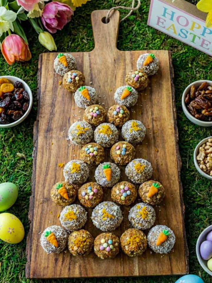 Top view of mini carrot cake energy bites arranged in a bunny shape on a wooden serving board, decorated with coconut coating, sprinkles, and tiny carrot toppers with Easter decor around.