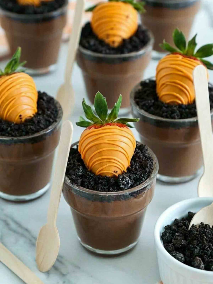 Clear dessert cups filled with chocolate pudding and topped with crushed cookie “dirt,” each decorated with a strawberry dipped in orange chocolate to resemble a carrot planted in soil, arranged on a white marble surface with wooden tasting spoons and extra cookie crumbs nearby.