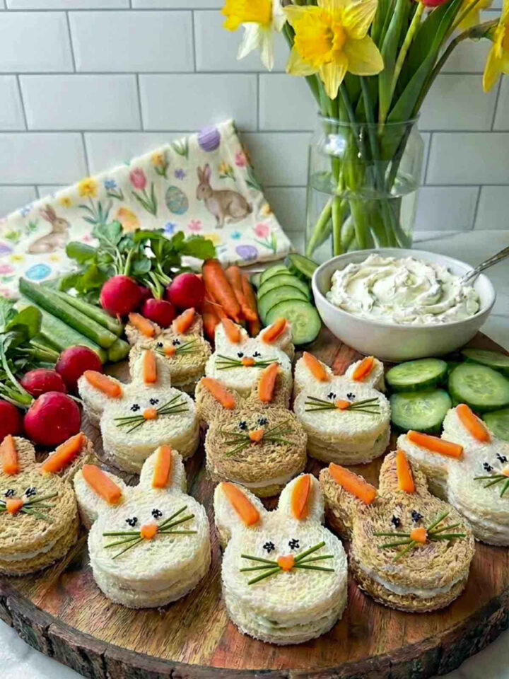 Mini bunny-shaped cream cheese sandwiches decorated with carrot ears, carrot nose, herb whiskers, and tiny black sesame eyes arranged on a rustic wooden board with fresh vegetables, cucumber slices, and a bowl of cream cheese, with spring flowers in the background.