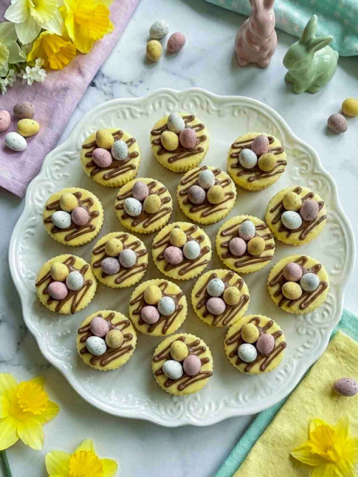 Mini no bake Easter cheesecake bites topped with chocolate drizzle and pastel candy eggs on a white plate with spring flowers and Easter decor.