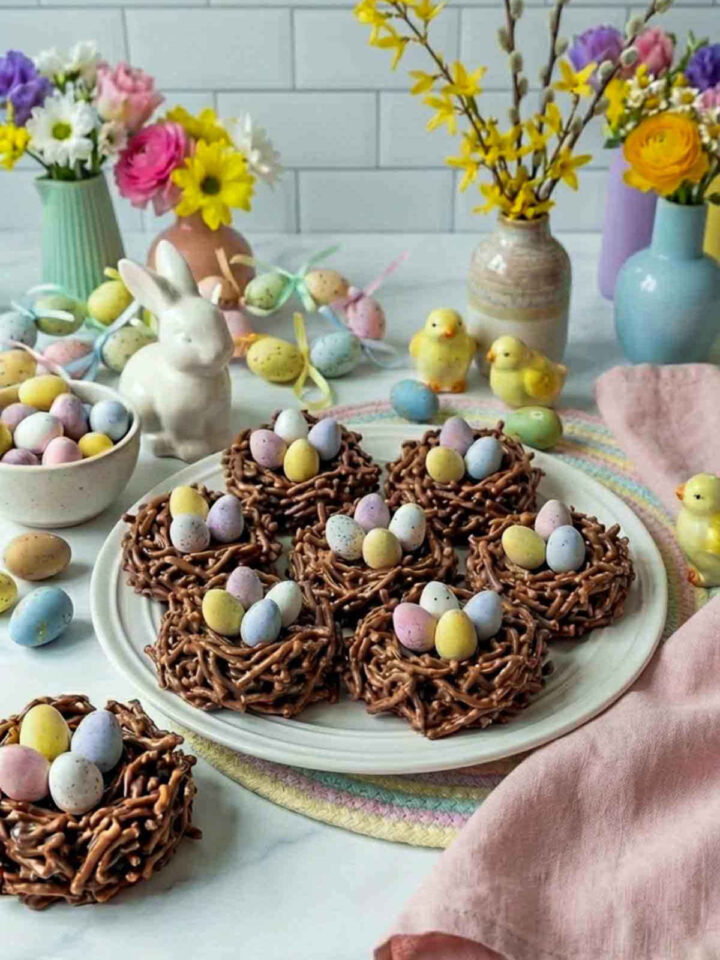 Chocolate no-bake Easter nest cookies made from chocolate-coated chow mein noodles shaped into small nests and filled with pastel candy chocolate eggs, arranged on a white plate with Easter decorations, flowers, and spring props in the background.