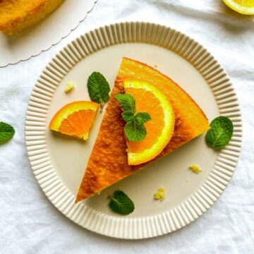 Slice of citrus cake topped with orange and lemon slices and fresh mint, served on a beige plate with crumbs and zest scattered around.