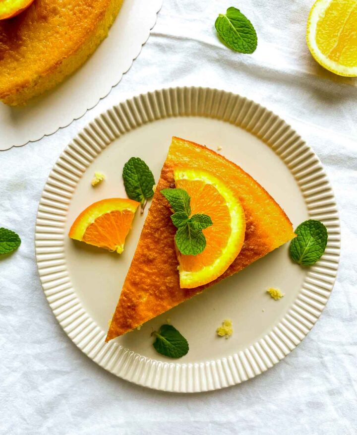 Top view of a citrus cake with one slice removed, and a plated slice topped with an orange wedge, with lemon halves and crumbs scattered on a light cloth background.