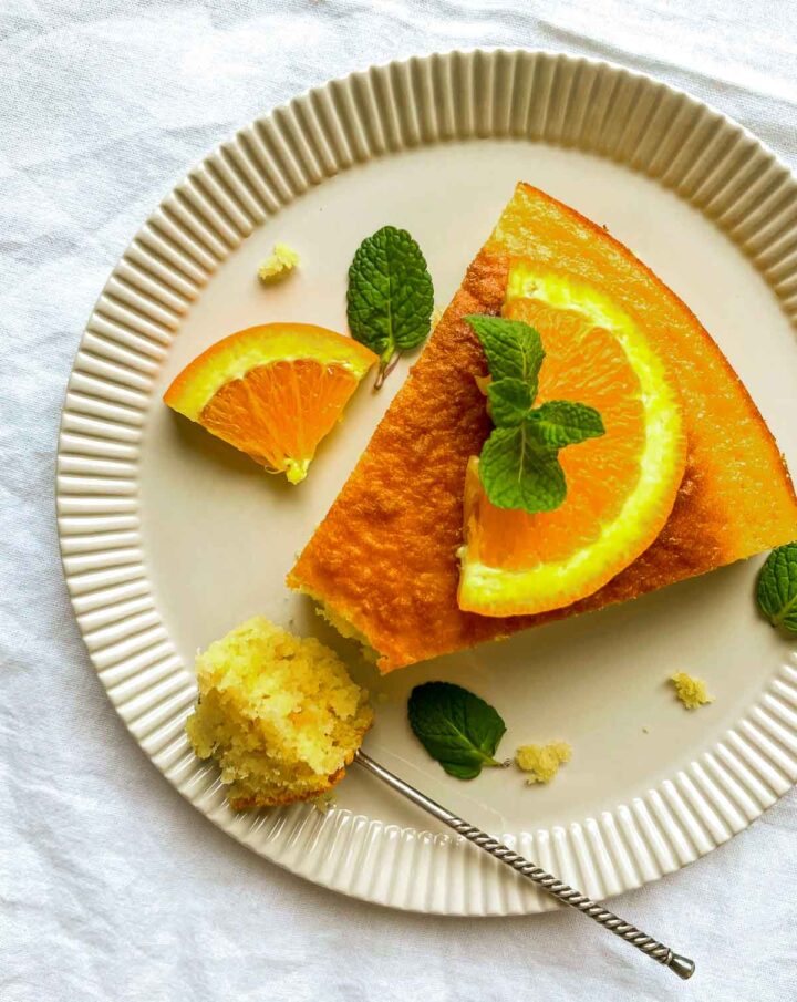 Slice of orange lemon cake garnished with citrus wedges and mint on a ridged beige plate, with a bite taken out and a fork resting beside it.