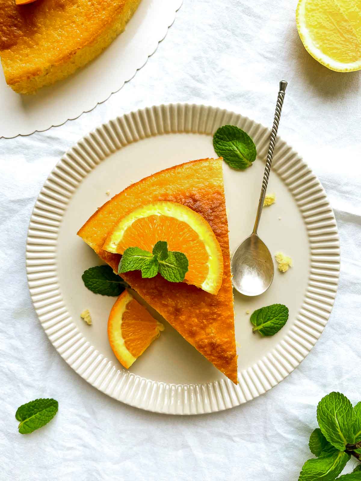 Top view of a citrus cake with one slice removed, and a plated slice topped with an orange wedge, with lemon halves and a spoon.