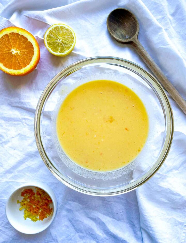 Glass mixing bowl filled with smooth citrus batter, surrounded by halved orange and lemon, zest in a small dish, and a wooden spoon on a white cloth.