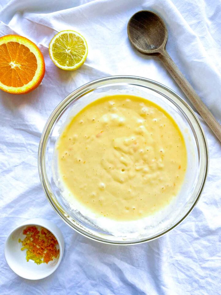 Glass bowl with slightly thickened citrus cake batter showing small lumps, surrounded by halved orange and lemon, zest in a small dish, and a wooden spoon on white fabric.