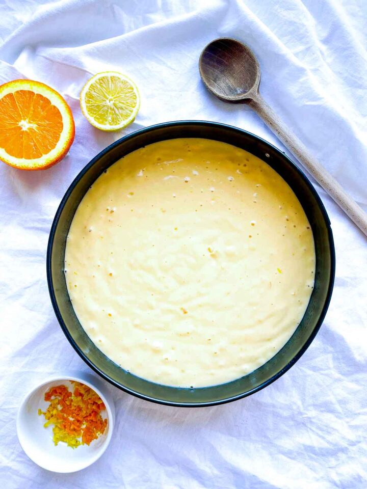 Citrus cake batter poured into a round baking pan, with halved orange and lemon, zest in a small dish, and a wooden spoon on white fabric.