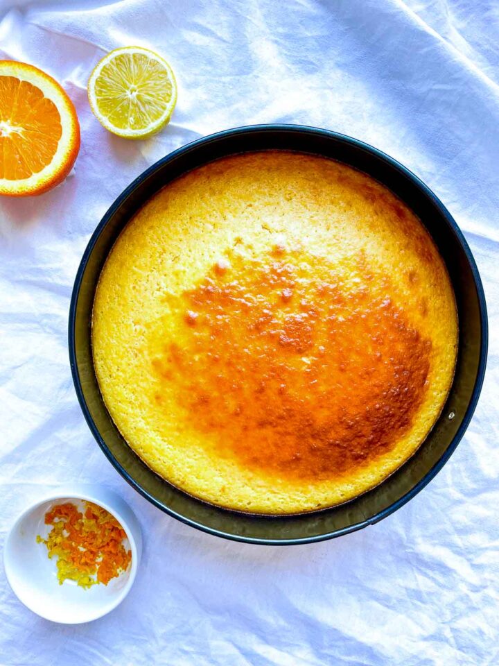Freshly baked citrus cake in a round pan with a golden brown top, surrounded by halved orange and lemon and a small dish of zest on white fabric.