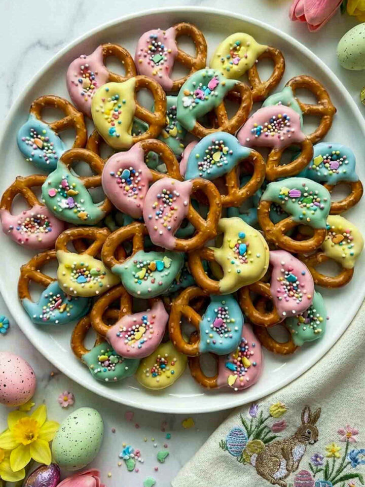 Pastel chocolate dipped pretzels with colorful Easter sprinkles arranged on a white plate with spring flowers and pastel Easter eggs.