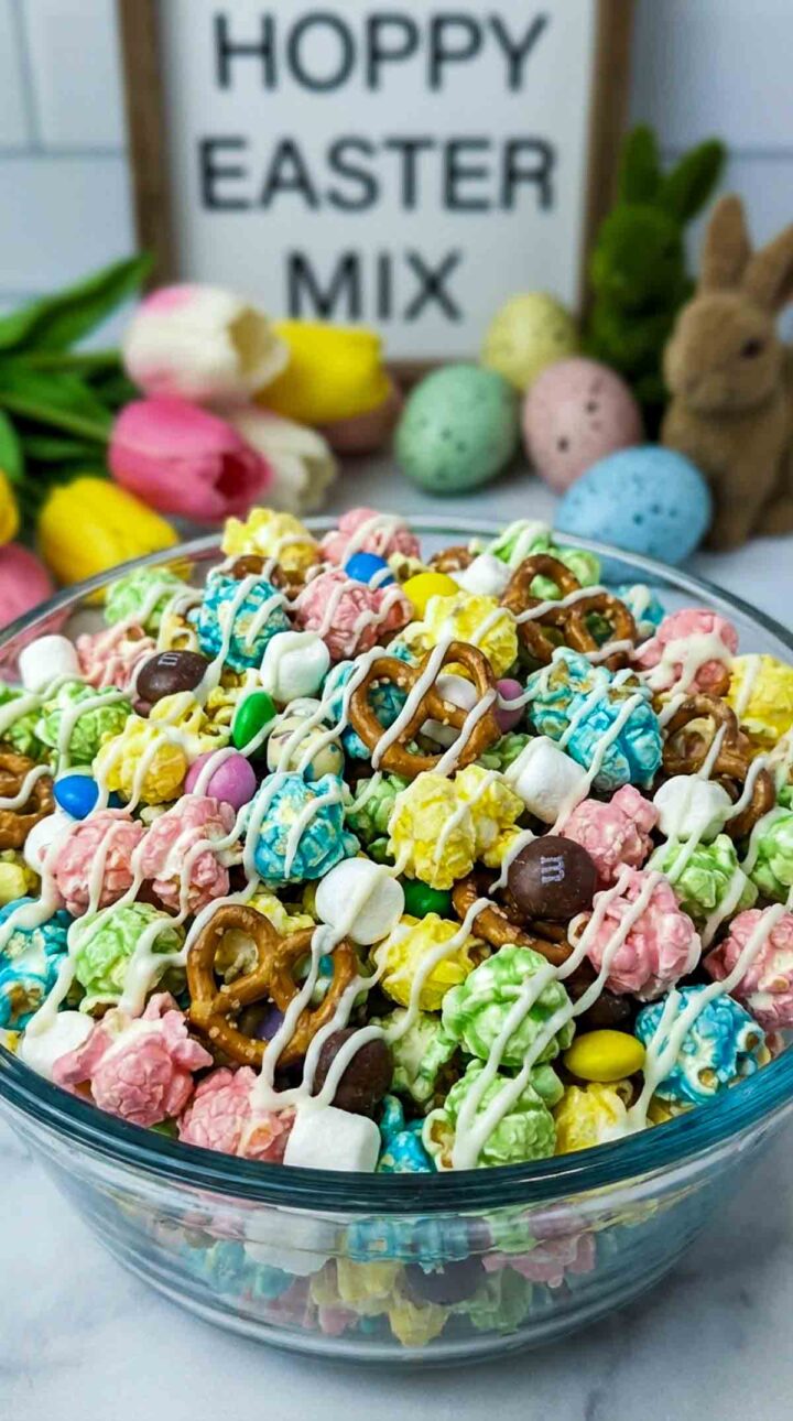 Large glass bowl filled with pastel-colored popcorn coated in yellow, pink, blue, and green candy melts, mixed with mini pretzels, mini marshmallows, and candy-coated chocolates, lightly drizzled with white chocolate, set on a white marble surface with tulips, pastel Easter eggs, and a “Hoppy Easter Mix” sign in the background.