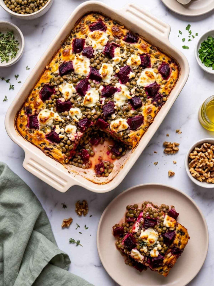 A cream ceramic baking dish filled with a roasted beet and lentil casserole topped with melted goat cheese and thyme, with one square removed and plated below showing whole lentils and beet cubes inside, surrounded by bowls of lentils, roasted beets, goat cheese, walnuts, and herbs on a marble surface.