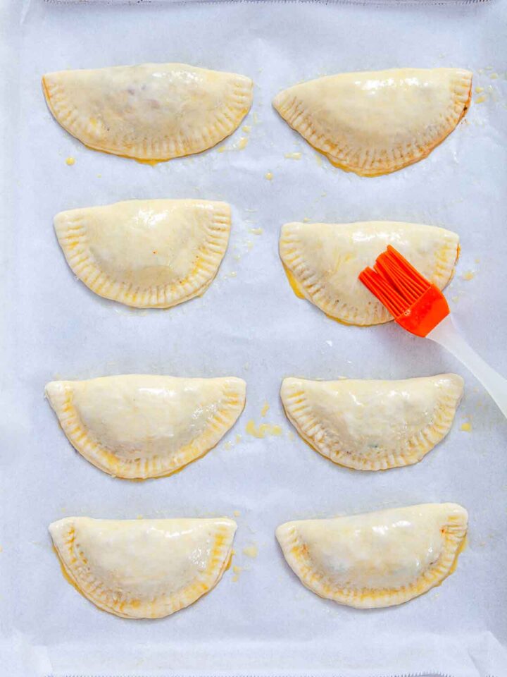 Unbaked sausage pizza pockets on a baking sheet being brushed with egg wash.