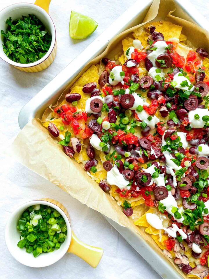 Sheet pan nachos topped with melted cheese, red kidney beans, salsa, sliced olives, sour cream, and chopped green onions, served with lime wedges and fresh herbs.